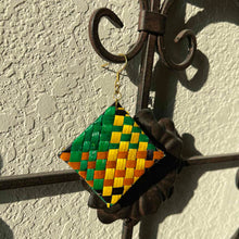 Load image into Gallery viewer, Decorative woven square ornament with green, yellow, and brown pattern hanging on a metal hook against a textured wall.