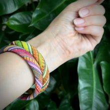 Load image into Gallery viewer, Hand wearing a colorful bracelet with green leaves in the background