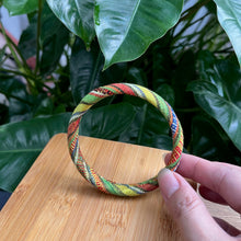 Load image into Gallery viewer, Hand holding a colorful braided bracelet with green leaves in the background
