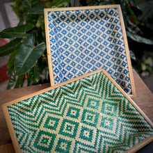 Load image into Gallery viewer, Two woven trays with geometric patterns on a wooden surface with plants in the background.
