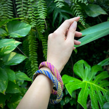 Load image into Gallery viewer, Hand wearing a colorful bracelet with green leaves in the background
